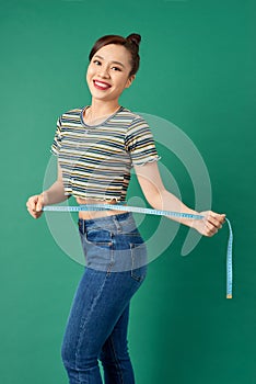 Beautiful young Asian woman measuring her waist with a measure tape over green background