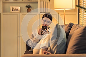 a beautiful young asian woman is pictured using her smartphone while comfortably leaning back on a couch in her cozy, warmly lit