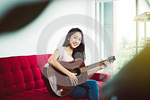 Beautiful young asian woman playing guitar while sitting on couch in living room at home