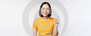 Beautiful young asian woman pointing finger up, smiling and looking amused at camera, showing advertisement