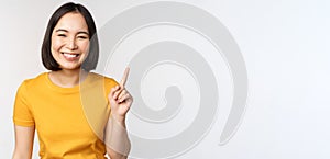 Beautiful young asian woman pointing finger up, smiling and looking amused at camera, showing advertisement