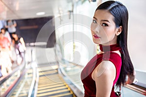 Beautiful young asian woman in red dress going down the escalator in shopping mall . happy elegant lady . copy space