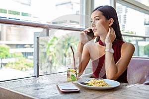 Beautiful young asian woman in red dress sitting in the restaurant looking out the window calling with smartphone . happy elegant