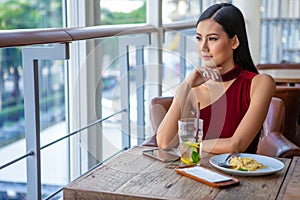 Beautiful young asian woman in red dress sitting in the restaurant looking out the window . happy elegant lady sitting on table