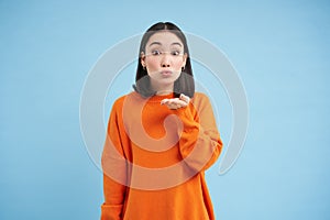 Beautiful young asian woman sends air kiss on her palm, kissing you, standing over blue background