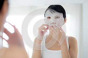 Beautiful young asian woman with sheet facial mask and looking mirror at home, beauty girl applying cosmetic and makeup.