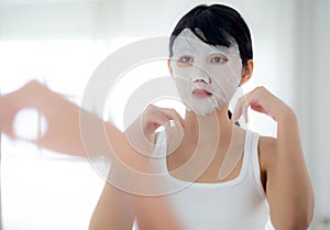 Beautiful young asian woman with sheet facial mask and looking mirror at home, beauty girl applying cosmetic and makeup.