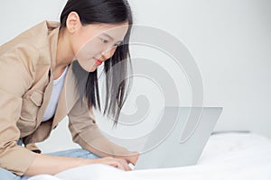 Beautiful young asian woman sitting on bed using laptop computer working from home in the bedroom.