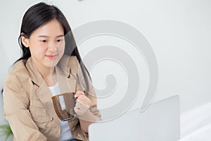 Beautiful young asian woman sitting on bed using laptop computer working from home and holding coffee cup.
