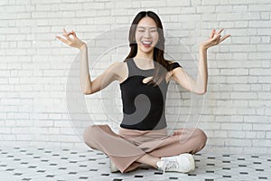 Beautiful young Asian woman sitting in lotus position meditate mudra fingers and laughing on white brick wall background. Healthy