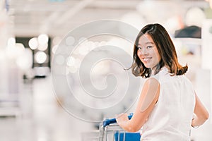 Beautiful young Asian woman smiling, with shopping cart, shopping center or department store scene, blur bokeh background