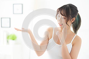 Beautiful young asian woman smiling and showing skin care product for advertisement at bedroom