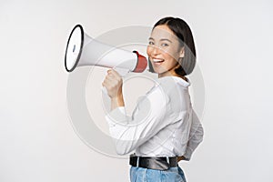 Beautiful young asian woman talking in megaphone, screams in speakerphone and smiling, making announcement, shout out