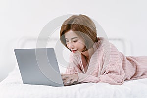 Beautiful young Asian woman in warm knitted pink clothes lying on bed while using laptop computer in bedroom. Lifestyle, comfort
