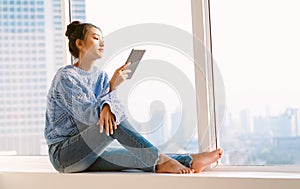 Beautiful young Asian woman in warm knitted sweater and using tablet computer while sitting on a window sill at home. Lifestyle,