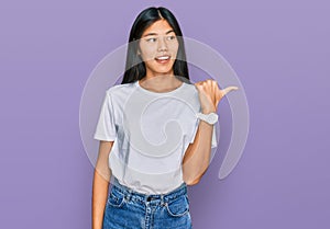 Beautiful young asian woman wearing casual white t shirt smiling with happy face looking and pointing to the side with thumb up