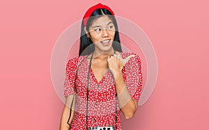 Beautiful young asian woman wearing dress and vintage camera smiling with happy face looking and pointing to the side with thumb