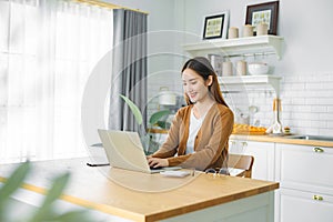 Beautiful young Asian woman working with laptop computer in home kitchen