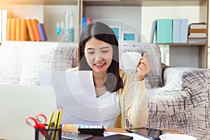 Beautiful young asian woman working on laptop computer and paperwork while sitting at living room, drinking hot coffee. Asia