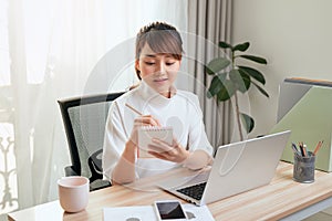 Beautiful young Asian woman writing notebook while working with laptop at home