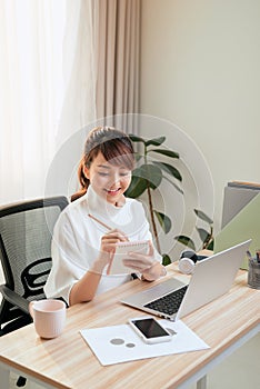 Beautiful young Asian woman writing notebook while working with laptop at home