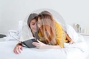 Beautiful young asian women LGBT lesbian happy couple reading book lying together in bed under blanket at home.