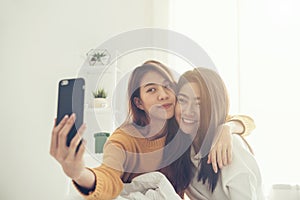 Beautiful young asian women LGBT lesbian happy couple sitting on bed hug and using phone taking selfie together bedroom at home.