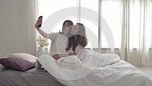 Beautiful young asian women LGBT lesbian happy couple sitting on bed hug and using phone taking selfie together bedroom at home.