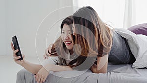 Beautiful young asian women LGBT lesbian happy couple sitting on bed hug and using phone taking selfie together bedroom at home.
