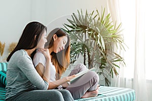 Beautiful young asian women LGBT lesbian happy couple sitting on sofa reading book together near window in living room at home.