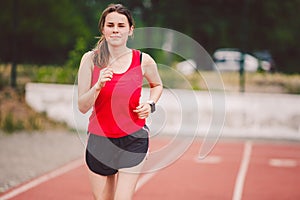 Beautiful young athlete Caucasian woman with big breasts in red T-shirt and short shorts jogging, running in the stadium with red