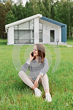 Beautiful young attractive woman sitting on green lawn with modern house on background talking on cell phone looking away with