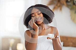 Beautiful young black woman applying face cream from jar, wearing bath towel, enjoying spa procedure at home