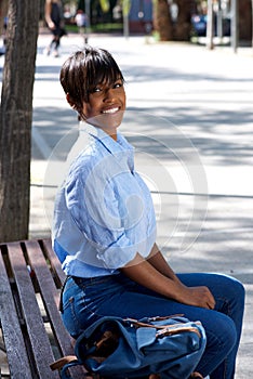 Beautiful young black woman sitting on bench outside with bag