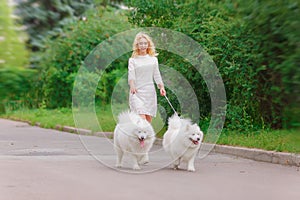 Beautiful young blond girl in dress walking with two white fluffy dogs in summer garden