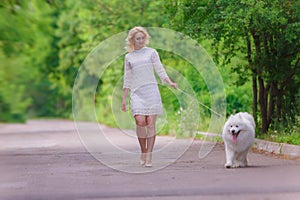 Beautiful young blond girl in dress walking with a white fluffy dog in summer garden