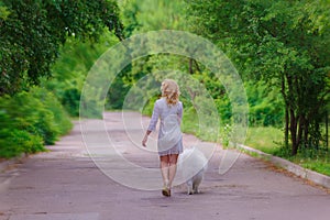 Beautiful young blond girl in dress walking with a white fluffy dog in summer garden