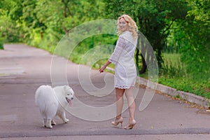 Beautiful young blond girl in dress walking with a white fluffy dog in summer garden