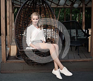 Beautiful young blond girl with long legs sitting in a wicker chair at an outdoor cafe on a warm summer evening, smiling and look