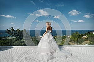 Beautiful young blond model girl in white dress standing with back to the camera and looking at sea