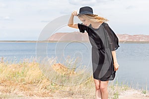 Beautiful young blond woman in a black dress and a light black hat in the desert and the wind blowing her hair in a hot summer day