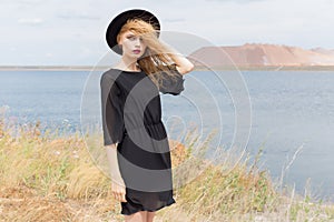 Beautiful young blond woman in a black dress and a light black hat in the desert and the wind blowing her hair in a hot summer day