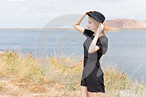 Beautiful young blond woman in a black dress and a light black hat in the desert and the wind blowing her hair in a hot summer day