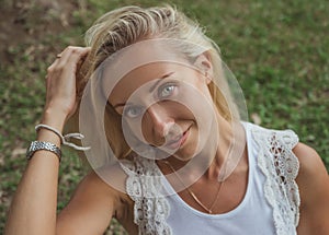 Beautiful young blond woman sits on the green grass and looks at the camera with beautiful gray eyes. Modern lifestyle