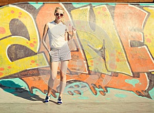 Beautiful young blond woman in sunglasses and a lollipop stands on graffiti background. Toned in warm colors