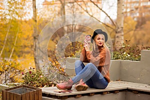 Beautiful young blonde in a brown warm sweater, black felt hat, blue jeans and boots sits on a bench in the autumn in the park,