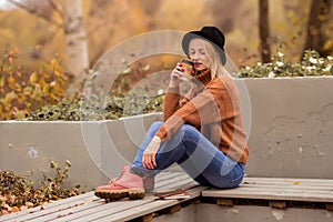Beautiful young blonde in a brown warm sweater, black felt hat, blue jeans and boots sits on a bench in the autumn in the park,