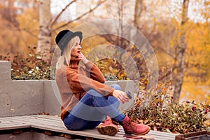 Beautiful young blonde in a brown warm sweater, black felt hat, blue jeans and boots sits on a bench in the autumn in the park,