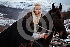 Beautiful young blonde on a crow. Woman viking with a black horse against the background of mountains