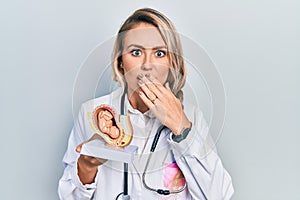 Beautiful young blonde doctor woman holding anatomical model of female uterus with fetus covering mouth with hand, shocked and
